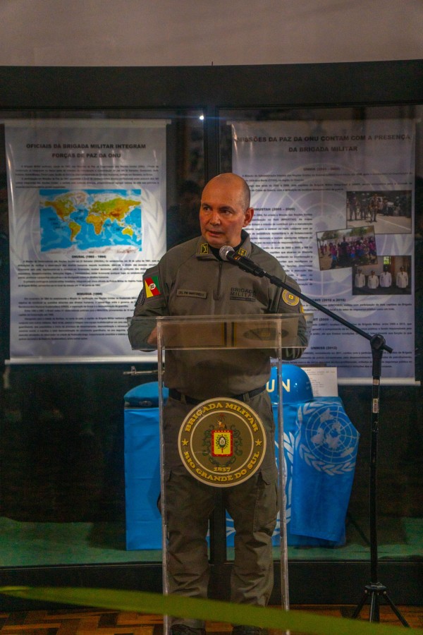 Fotografia colorida mostra o coronel PM Álvaro Martinelli discursando em um púlpito com o brasão da Brigada Militar. Ao fundo aparecem painéis expositivos relacionados à participação de policiais militares em missões de paz da ONU.