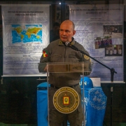 Fotografia colorida mostra o coronel PM Álvaro Martinelli discursando em um púlpito com o brasão da Brigada Militar. Ao fundo aparecem painéis expositivos relacionados à participação de policiais militares em missões de paz da ONU.