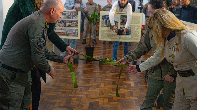 Fotografia colorida registra o momento do descerramento da fita inaugural da exposi&ccedil;&atilde;o. Autoridades civis e militares seguram uma fita verde enquanto realizam a abertura oficial da mostra. Ao fundo aparecem convidados, pain&eacute;is hist&oacute;ricos e o ambiente interno do museu.