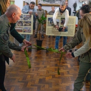 Fotografia colorida registra o momento do descerramento da fita inaugural da exposi&ccedil;&atilde;o. Autoridades civis e militares seguram uma fita verde enquanto realizam a abertura oficial da mostra. Ao fundo aparecem convidados, pain&eacute;is hist&oacute;ricos e o ambiente interno do museu.