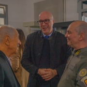 Fotografia colorida mostra três homens conversando durante a abertura da exposição. Ao centro está o presidente da Associação Amigos do Museu da Brigada Militar, coronel da reserva remunerada Jerônimo Carlos Santos Braga. À direita, o coronel PM Álvaro Martinelli, chefe do Estado-Maior da Brigada Militar. Ao fundo, visitantes observam o ambiente do museu.