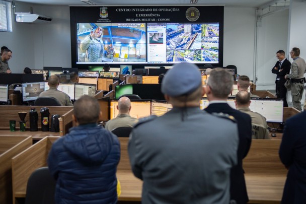 Participantes observam demonstração do sistema da Brigada Militar no COPOM, com imagens exibidas em telão.