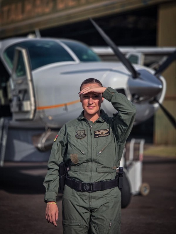 A PM aparece sob o sol, fardada e à frente de uma aeronave da Instituição. 