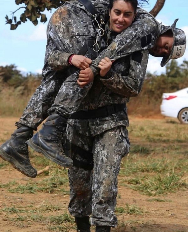 A PM aparece carregando um colega durante uma um exercício simulado de Atendimento Pré-Hospitalar em Combate (APH-C). O local é um campo aberto. 