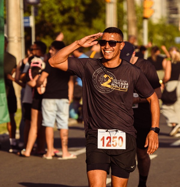 Soldado PM aparece prestando continência enquanto participa da corrida. Ele veste a camiseta da prova e está sob o número 1250. O evento aconteceu em dia ensolarado e é possível ver o público em volta. 