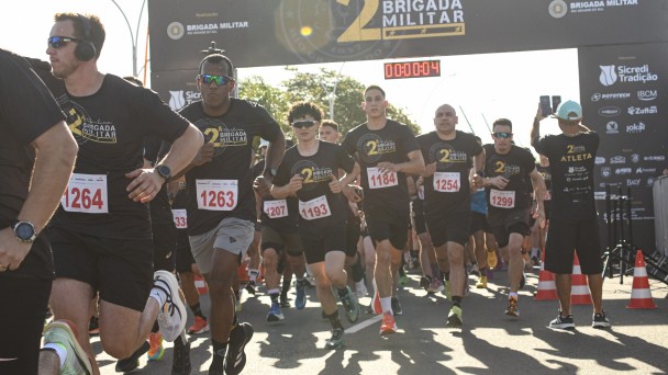 Imagem mostra o momento de in&iacute;cio do percurso. Aparecem os participantes correndo vestindo as camisetas da prova. Ao fundo, aparece o p&oacute;rtico com cron&ocirc;metro da corrida. 