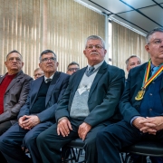 A imagem mostra antigos diretores do Departamento de Pessoal e Finanças sentados na primeira fila do auditório. Os homenageados vestem trajes sociais e acompanham a cerimônia com atenção.