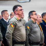 A imagem registra autoridades, militares estaduais e convidados em posição de respeito durante a execução de ato solene. Os participantes estão alinhados no auditório, voltados para a área central da cerimônia.