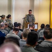 A imagem mostra o diretor do Departamento de Pessoal e Finanças, coronel PM Cléber Rodrigues dos Santos, em pé atrás de um púlpito realizando seu pronunciamento. Militares estaduais acompanham a fala sentados no auditório.