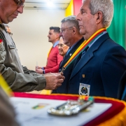A imagem mostra o momento da entrega da Comenda do Departamento de Pessoal e Finanças. Um oficial da Brigada Militar coloca a medalha no pescoço de um homenageado vestido com traje social. Ao fundo, outras autoridades aguardam a homenagem.