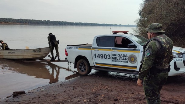 PMs do Comando de Pol&iacute;cia Ambiental est&atilde;o junto a uma viatura na beira de um curso d'&aacute;gua onde est&aacute; um barco com um PM dentro. 