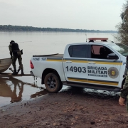 PMs do Comando de Pol&iacute;cia Ambiental est&atilde;o junto a uma viatura na beira de um curso d'&aacute;gua onde est&aacute; um barco com um PM dentro. 