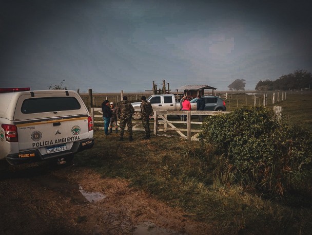 PMs aparecem na porteira de uma propriedade rural em diálogo com moradores. A viatura aparece à esquerda e dois veículos estão do lado de dentro do endereço. 