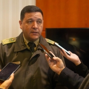 Oficial da Brigada Militar concede entrevista em ambiente interno. Jornalistas seguram celulares e microfones próximos a ele.