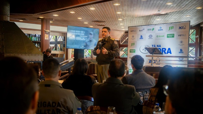 Oficial da Brigada Militar fala ao p&uacute;blico durante palestra, segurando microfone, em sal&atilde;o com participantes sentados e painel do T&aacute; na Hora ao fundo.