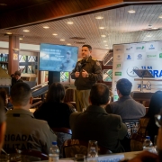 Oficial da Brigada Militar fala ao p&uacute;blico durante palestra, segurando microfone, em sal&atilde;o com participantes sentados e painel do T&aacute; na Hora ao fundo.